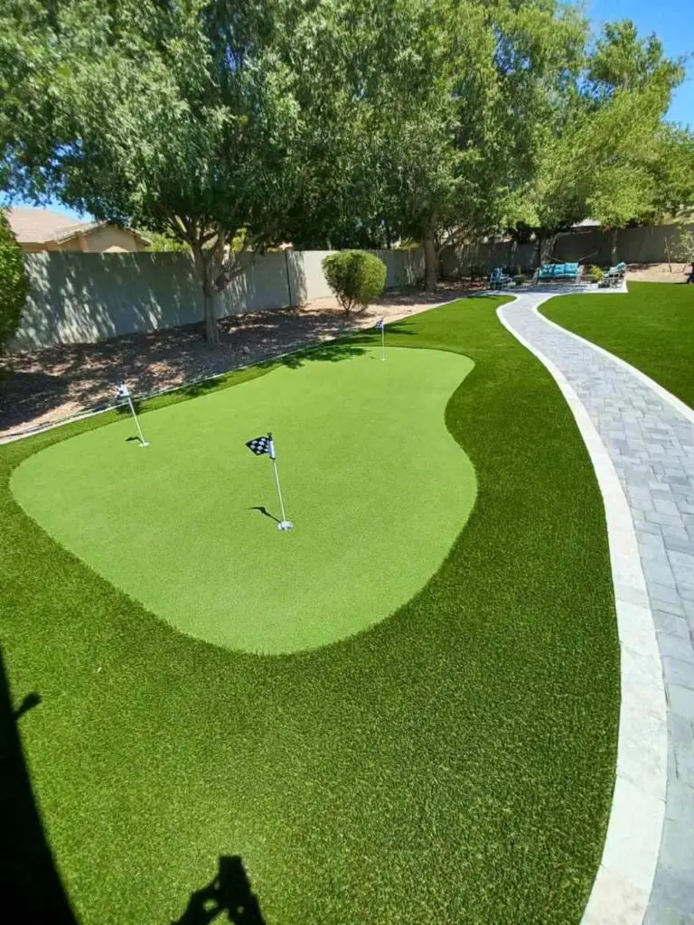 A backyard golf putting green with three flags is surrounded by neatly trimmed grass and bordered by a stone pathway. Trees and patio furniture are visible in the background under a clear blue sky.